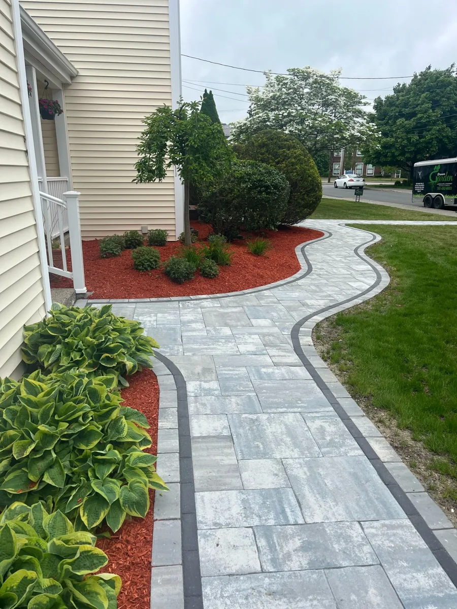Gray paver walkway curving past planted beds beside a home